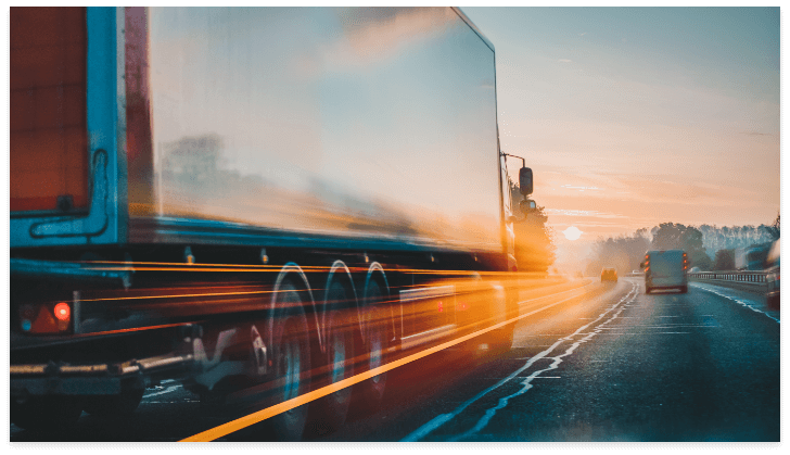 A motion-blurred truck speeds down a wet highway at sunrise, light trails emphasizing its urgency for holiday shipping. Another vehicle is visible ahead under an orange sky, highlighting the rush to meet Holiday Shipping Deadlines.
