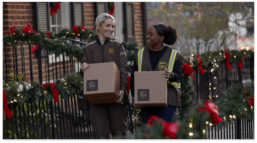 Two UPS workers holding packages and smiling at each other while walking outside a building decorated with holiday garlands and red bows, ready to meet Holiday Shipping Deadlines.
