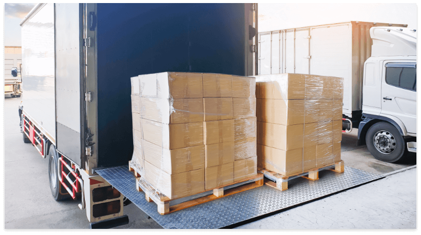 Two pallets loaded with stacked cardboard boxes, wrapped in clear plastic, sit on the hydraulic lift of a delivery truck at a loading dock, ready to meet holiday shipping deadlines. Another white truck is visible in the background.