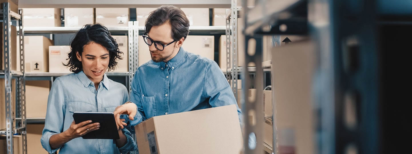 while the woman is pointing at a tablet. Both are wearing casual blue shirts and appear to be discussing something on the screen.|Two people in denim shirts stand in a storage area with shelves. One holds a box while the other points at a tablet screen