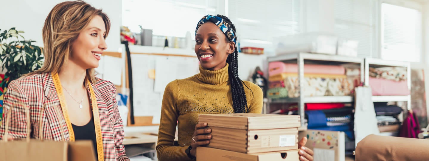 the other in a plaid jacket holds the tablet. A green circle with a white financial icon is on the left.|Two women smile while looking at a tablet in a workspace. One leans on a stack of boxes