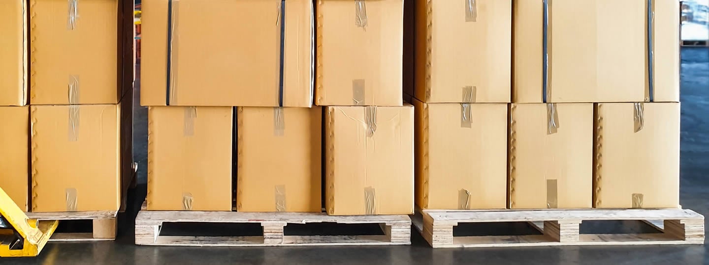 while a blue overlay graces the right side of the image.|A manual pallet jack with yellow wheels stands beside stacks of brown cardboard boxes on wooden pallets