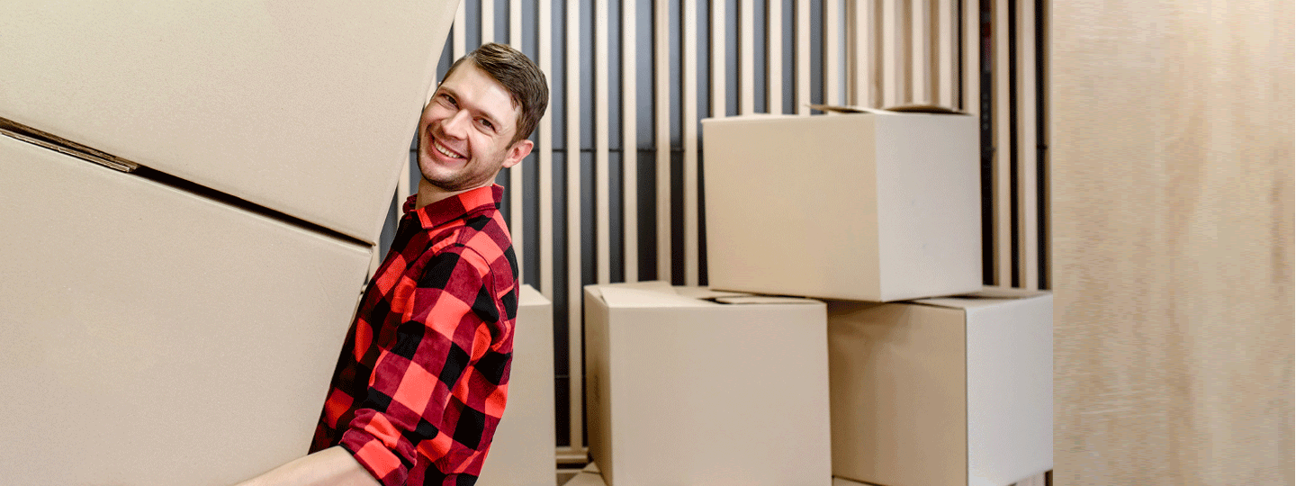 A smiling man in a red plaid shirt carries an oversized package and another large box, with more boxes stacked in the background.