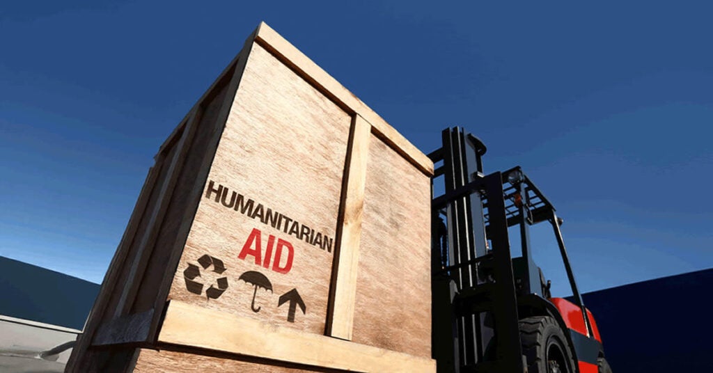 A forklift lifts a large wooden crate labeled HUMANITARIAN AID, marked with recycling, fragile, and upward symbols—an ideal case study in efficient logistics set against a clear blue sky.