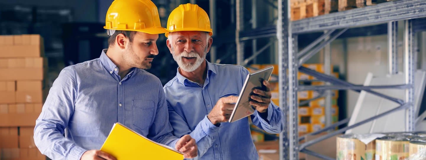 while the other uses a tablet. Shelves with boxes loom in the background as they discuss or review details.|Two men in yellow hard hats stand in a warehouse