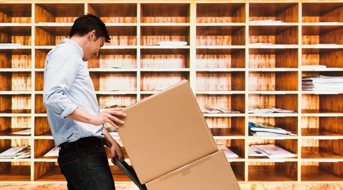 A delivery person in a brown uniform pushes a dolly with large cardboard boxes in front of a striped background. On the left side