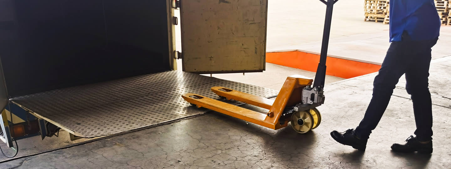 and the floor has directional arrows marked. The person is wearing dark pants and shoes.|A person in a blue jacket is using a manual pallet jack to unload rejected freight from a truck. The trucks rear door is open