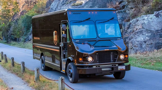 A person in a blue shirt is unloading boxes from the open back of a white van onto a flat surface. Various packages are scattered around