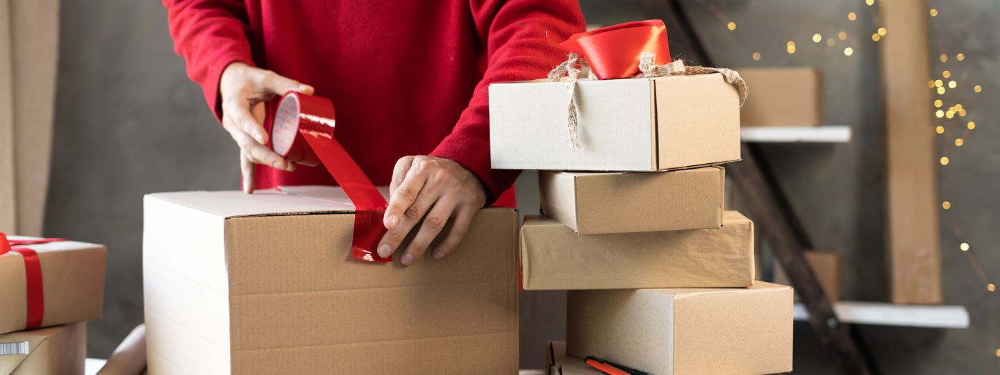 a person diligently seals a cardboard box with red tape on a table. Surrounding them are more boxes and packing materials