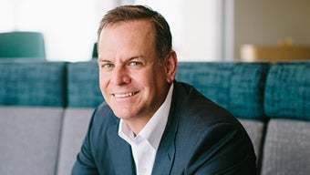 A smiling man in a business suit sits on a gray and teal cushioned bench