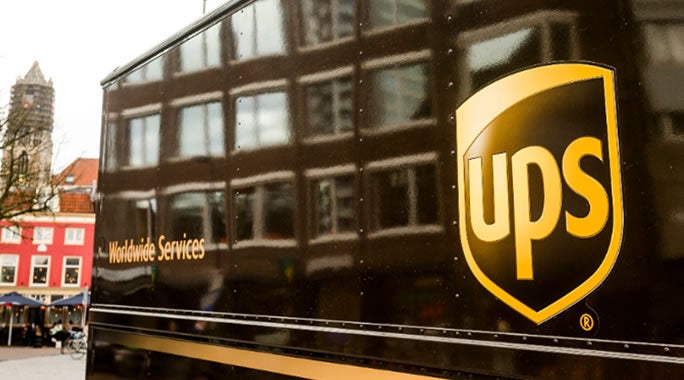 A delivery person in a uniform hands a package to a person in a maroon sweater at a doorway. Theyre smiling