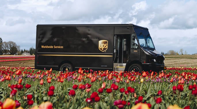 UPS logo on the side of a dark delivery truck, with a pattern of blue and white arrows pointing right on the left side of the image, highlighting a swift UPS Pickup service.