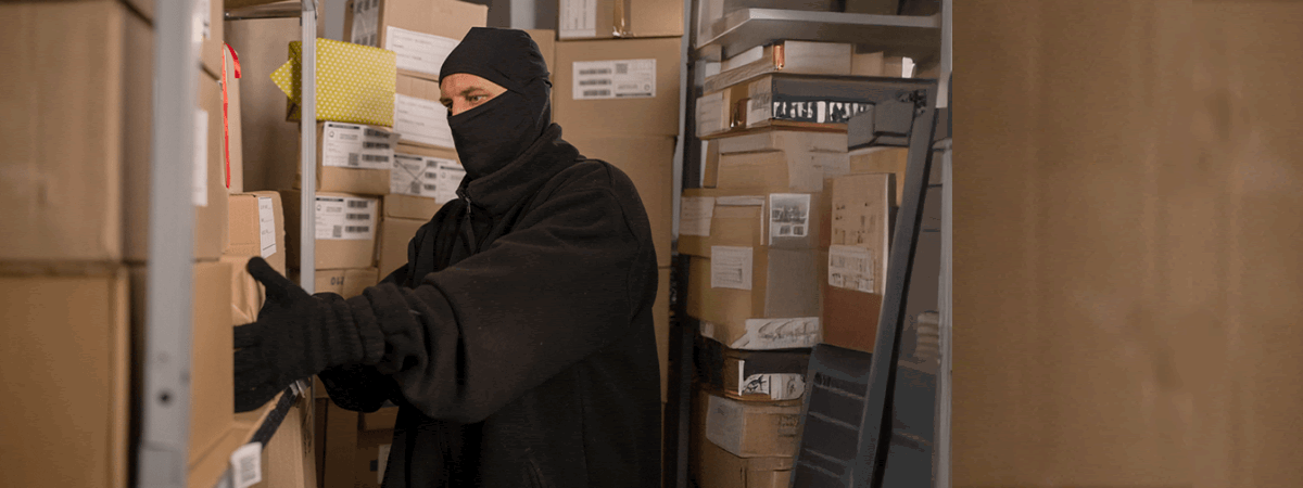 A person wearing a black mask, gloves, and dark clothing is surrounded by shelves stacked with cardboard boxes, appearing to search through or take specialty freight packages in a storage room.