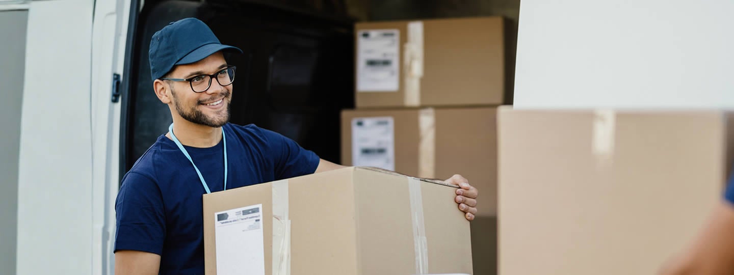 A smiling delivery worker wearing glasses and a blue cap unloads cardboard boxes from a van. Several labeled packages are stacked inside the vehicle, showcasing a careful White Glove Shipping process.