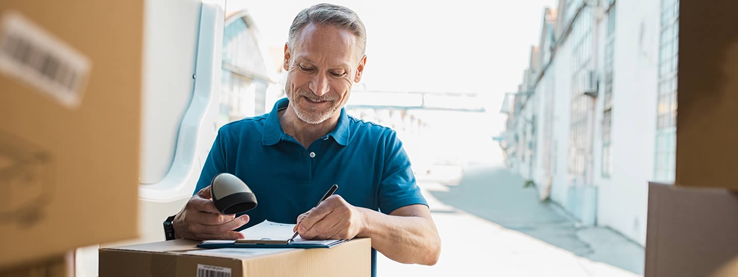 smiling as he signs a document on a clipboard. Surrounded by boxes