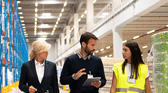 Three people walk through a warehouse. A man in the center holds a tablet