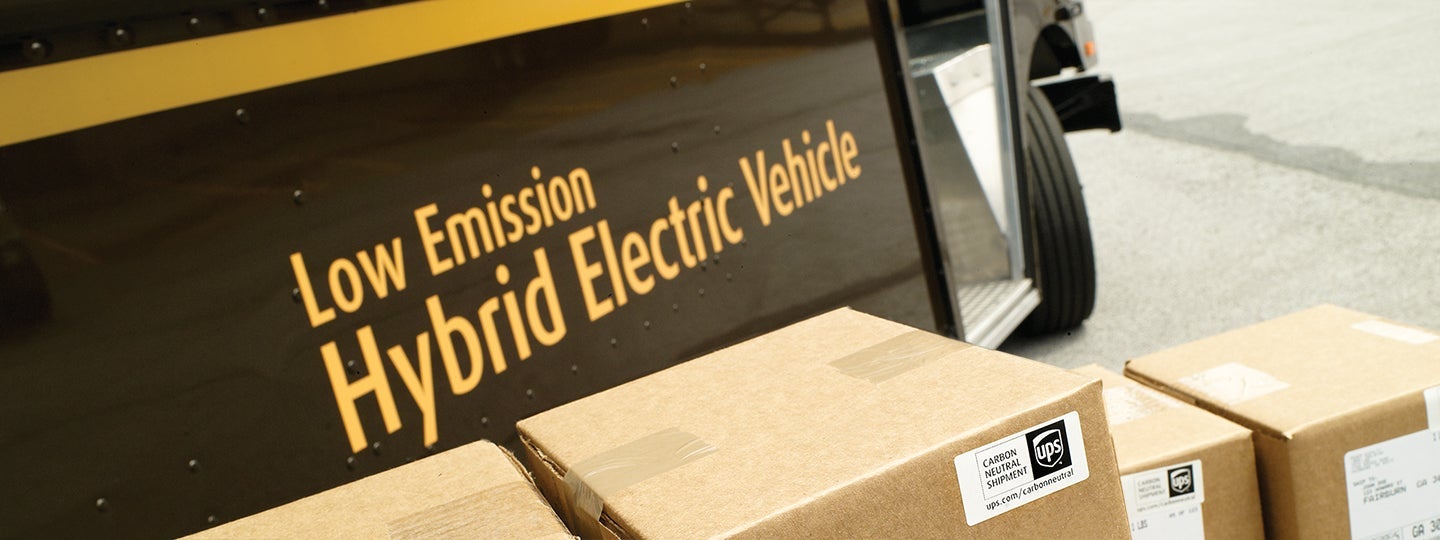 and part of a wheel is visible in the background.|Close-up of cardboard packages in front of a delivery truck with Low Emission Hybrid Electric Vehicle on its side