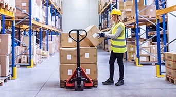 A woman in a warehouse wears a yellow hard hat and reflective vest as she operates a pallet jack