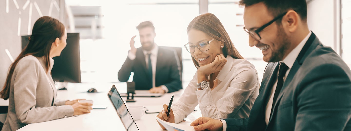while the man has short hair and a beard. Another person is blurred in the background.|In a bright office setting