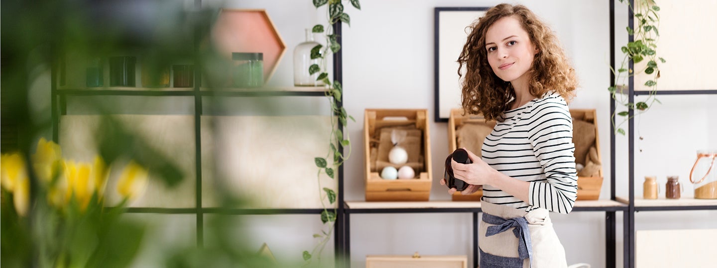 a person in a striped shirt and apron stands surrounded by shelves of wooden boxes and plants
