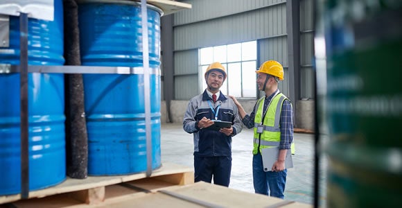 Two workers in safety gear stand in a warehouse, discussing energy logistics near stacked blue barrels. One holds a clipboard, the other gestures with a tablet. Industrial shelves and large windows are visible in the background.