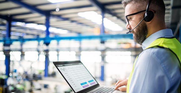 A man wearing a safety vest and headset uses a laptop in an industrial warehouse setting, overseeing energy logistics operations and monitoring data on the screen.