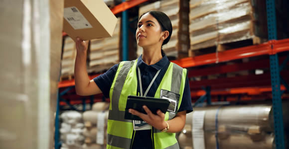 A warehouse worker in a yellow safety vest holds a box up to inspect it while carrying a tablet, standing among tall storage shelves stocked with goods—demonstrating efficient 3PL shipping operations.