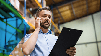 A man in a light blue shirt holds a clipboard and talks on the phone in a bustling small-business shipping hub. Shelves brimming with boxes fill the background