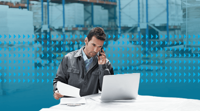 A man in a work jacket talks on the phone while looking at documents and a laptop in a warehouse with shelves and large pallets in the background.|A man in a warehouse sits at a desk with a laptop