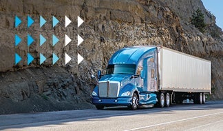 A blue semi-truck with a white trailer navigates the road beside a rocky cliff