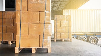 Stacks of cardboard boxes wrapped in plastic are neatly arranged on wooden pallets in the small-business-shipping-hub. Nearby