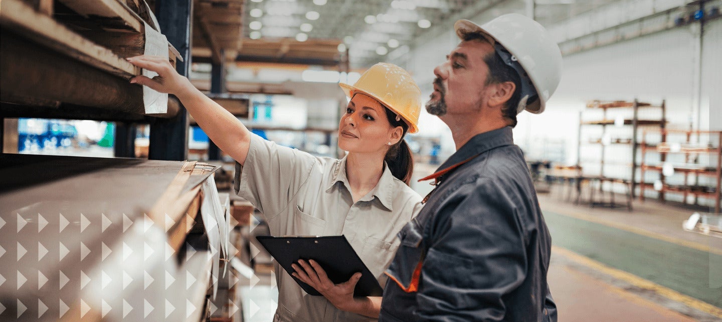 both in hard hats. One holds a clipboard while pointing at a shelf