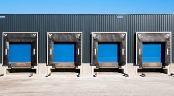Four loading docks with blue doors and black weather seals are evenly distributed against a gray industrial building under a clear blue sky. The concrete loading area in this small-business-shipping-hub is empty out front.|The image shows a row of five blue industrial loading dock doors set in a large black building