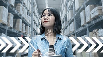 A person with glasses stands in a warehouse surrounded by tall stacks of boxes. They hold a tablet and appear focused. Overlaid icons represent technology and logistics