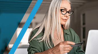 A woman with long white hair and glasses is holding a card while looking at a laptop screen. She is wearing a green shirt and is seated indoors. The image has a diagonal blue line on the left side.|A woman with long gray hair and glasses uses a laptop