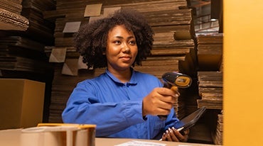 A woman with curly hair wearing a blue shirt is scanning a barcode with a handheld scanner in a warehouse filled with stacked cardboard boxes. She holds a tablet in her other hand.|A person in a blue uniform efficiently scans a package with a handheld scanner in the bustling warehouse