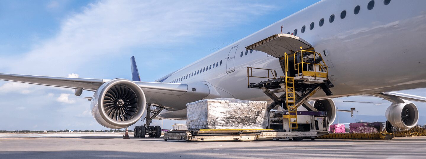 loading wrapped pallets onto the aircraft. The sky is clear with some clouds.|A large airplane on a tarmac with cargo being loaded into its side compartment. A worker operates a yellow lift to transport pallets wrapped in plastic. The sky is clear with a few clouds.