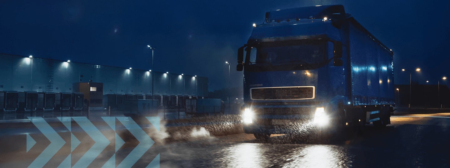 dimly lit industrial area at night. Its headlights illuminate the wet road. In the background