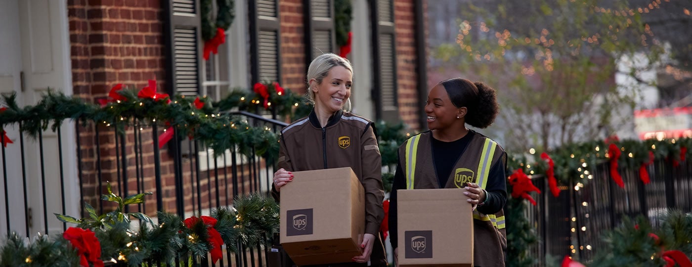 carry boxes adorned with UPS logos. Theyre walking along a decorated street with greenery and red bows