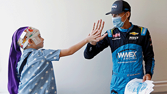 A child in a printed headscarf and pajamas high-fives a masked racecar driver from WWEX NASCAR