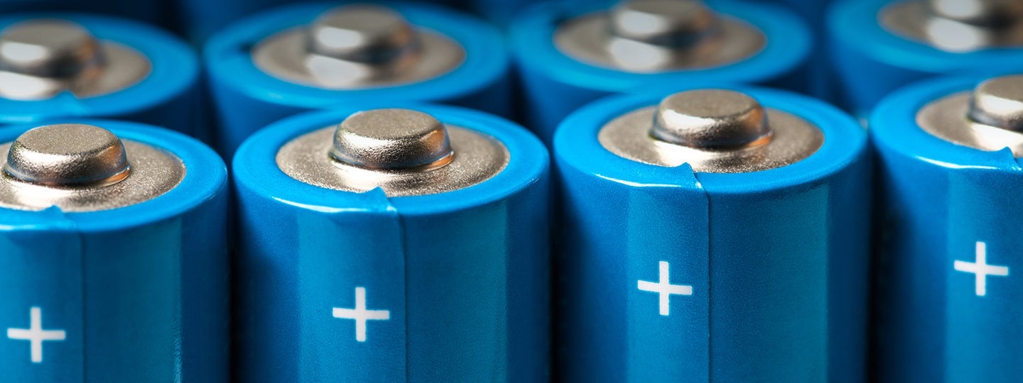 indicating the positive terminal. The image focuses on the battery tops in sharp detail.|Close-up of several blue cylindrical batteries standing upright in rows
