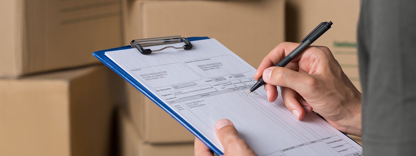 filling out a form with a pen. The background shows stacked cardboard boxes. Blue arrows are overlaid on the image