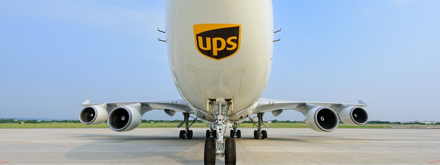with the UPS logo on the nose. To the left is a blue geometric shape featuring a white globe symbol. The sky is clear