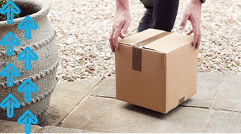 Person lowering a cardboard box onto a tiled surface beside a large plant pot. Blue arrows point upward on the left side of the image. Gravel surrounds the tiles.|A parcel carrier crouches down to lift a cardboard box placed on the paved surface. Gravel forms the background