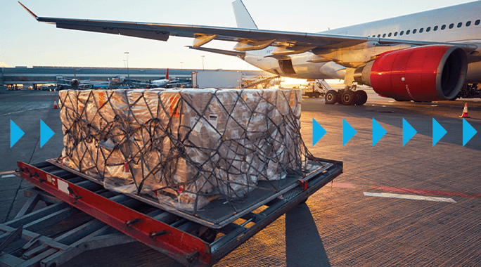 Cargo loaded on a dolly is ready for transportation in front of an airplane on a tarmac at sunset. Blue arrows indicate movement toward the aircraft.|As the sun casts long shadows across the tarmac