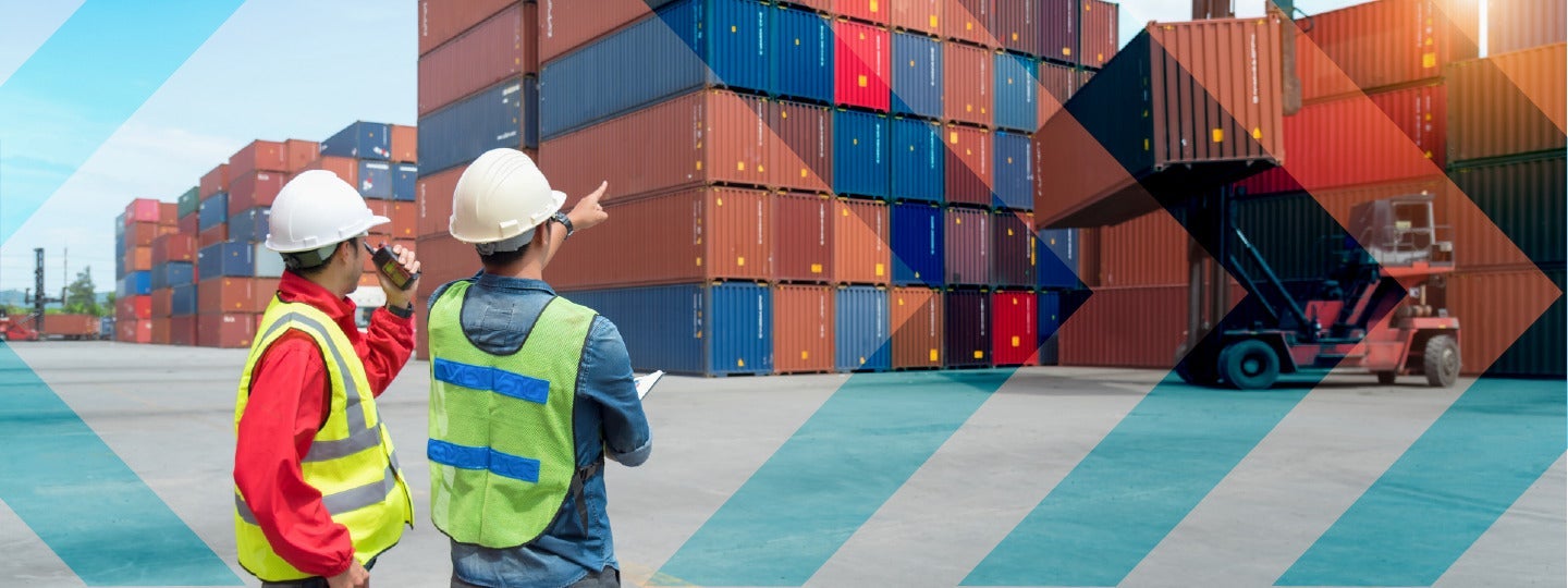overseeing the managed transportation process. One points towards a container being lifted by a crane in the busy shipping yard under a clear blue sky.|Two workers in hard hats and safety vests stand on a dock