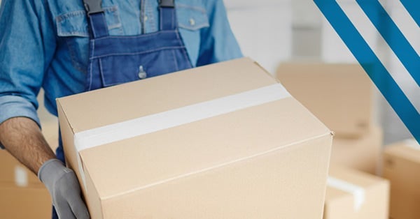 A person wearing a blue shirt and overalls carries a large cardboard box sealed with white tape. Gloved hands grip the box