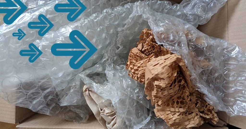 A cardboard box containing crumpled brown paper and clear bubble wrap. Blue arrow graphics are overlaid on the image