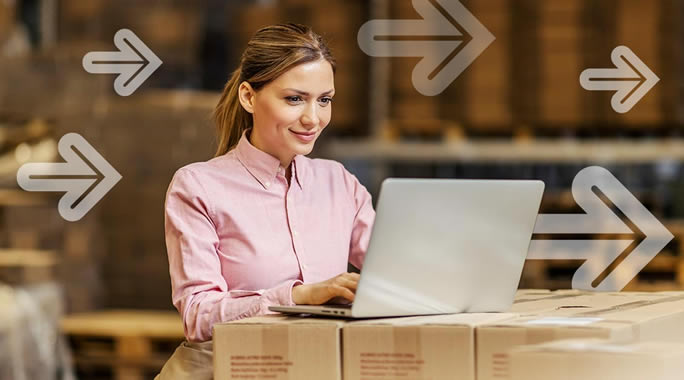 A woman in a pink shirt works on a laptop placed on stacked cardboard boxes in a warehouse. Transparent arrows pointing right are overlaid on the image