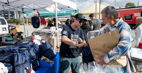 Two men stand at an outdoor Greater Good Charities donation event, one handing items to the other, who holds a large paper bag. Tables with bags, toys, and emergency shipping supplies are set up under tents in the background.